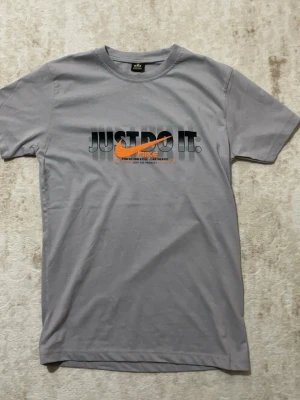 Grå Nike t-shirt med tryck - Snygg grå t-shirt från Nike med svart och orange tryck framtill där det står 'Just Do It' och en stor Nike-logga på ryggen. Klassisk passform och rund hals. Perfekt för dig som gillar sportig och stilren streetwear. Skickar samma dag vid köp. Passar dom som är 15 till 16 så storlek S ungefär 