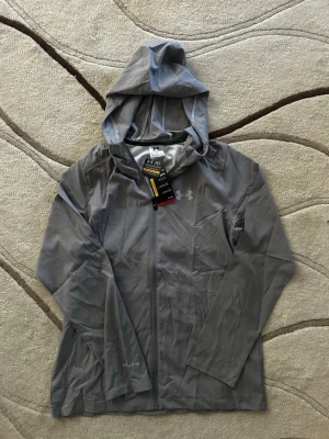 Grå vindjacka från Under Armour - Snygg grå vindjacka från Under Armour med huva och hel dragkedja framtill. Jackan har diskret logga på bröstet och stor logotyp på ryggen. Tillverkad i lätt och vattenavvisande material, perfekt för blåsiga dagar. Lång ärm och enkel, sportig design.