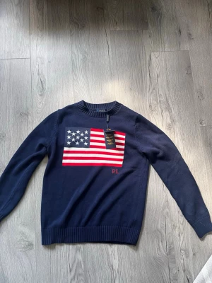  stickad tröja Ralph Lauren - Mörkblå stickad tröja från Polo Ralph Lauren med amerikansk flagga i rött, vitt och blått på bröstet. Rund halsringning och ribbade muddar. Klassisk och stilren design med RL-broderi nedtill på flaggan.