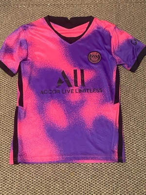 PSG matchtröja rosa/lila - Säljer en snygg PSG matchtröja i rosa och lila med svart logga och texten 'Accor Live Limitless' på bröstet. Tröjan har korta ärmar, V-ringning och är gjord i ett lätt och snabbtorkande material som passar perfekt till träning eller match.