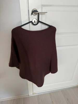 Mörkbrun ponchotröja - En stilren mörkbrun ponchotröja med vid passform och båtringad hals. Perfekt för lager på lager och ger en avslappnad men trendig look. Materialet ser mjukt och lite fluffigt ut, vilket gör den extra mysig under kyliga dagar.