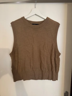 Beige/brun stickad väst  - Säljer en beige/brun stickad väst med ribbade kanter i hals, ärmhål och nederkant. Västen har en klassisk, rak modell och är perfekt att bära över en skjorta för en trendig lager-på-lager-look. Enkel och stilren design utan mönster eller extra detaljer.