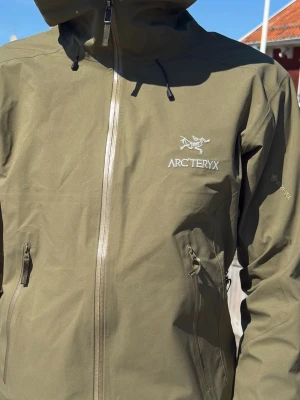 Olivgrön Arc'teryx regnjacka - Snygg olivgrön regnjacka från Arc'teryx med huva och vattentäta dragkedjor. Tillverkad i slitstarkt Gore-Tex-material som står emot regn och vind. Jackan har två praktiska sidofickor med dragkedja och justerbar huva. Perfekt för regning blåsigt eller bara en skön dag på havet🌊🎣🏄🏽‍♂️