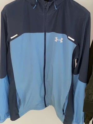 Under armour kofta - Snygg tvåfärgad vindjacka från Under Armour i mörkblått och ljusblått. Jackan har huva, hel dragkedja framtill, reflekterande detaljer och logga på bröstet samt stor logga på ryggen. Tillverkad i lätt och funktionellt material, perfekt för blåsiga dagar.