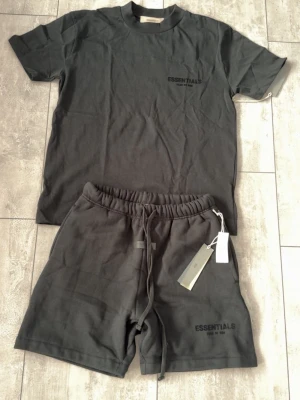 Svart set från Essentials (ÄKTA) - Säljer ett svart set från Essentials med t-shirt och shorts. T-shirten har rund hals och korta ärmar, medan shortsen har resår i midjan och dragsko. Båda delarna har diskret Essentials-logga tryckt framtill. Perfekt för en avslappnad och trendig look.