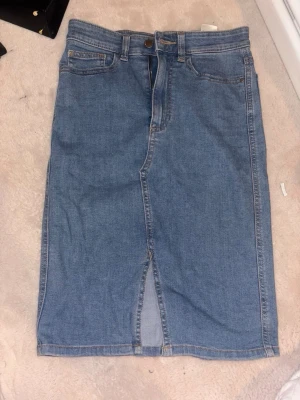 Blå jeanskjol med slits framtill - Säljer en klassisk blå jeanskjol i lång modell med slits framtill. Kjolen har femficksdesign, bälteshällor och stängs med knapp och dragkedja. Perfekt för dig som gillar en enkel och tidlös denimlook.