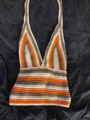 Randig virkad linnetopp i orange och beige - Säljer en virkad linnetopp med djup v-ringning och smala axelband. Toppen har ränder i orange, beige, grått och vitt och är croppad i modellen. Perfekt för sommaren och festivaler!