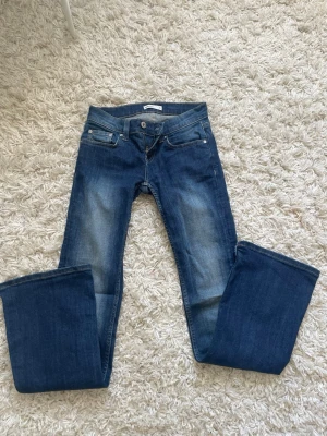 Blå bootcut jeans från Gina Tricot - Säljer ett par blå bootcut jeans från Gina Tricot med snygga slitningar framtill och dekorativa sömmar på bakfickorna. Klassisk femficksmodell med knapp och dragkedja. Perfekta till sneakers eller boots!