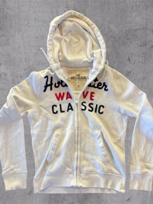 Vit hoodie från Hollister med tryck - Vit hoodie från Hollister med dragkedja framtill och broderad text i marinblått och rött på bröstet. Klassisk modell med två fickor fram och justerbar huva. Tillverkad i mjukt bomullsmaterial för en avslappnad look.