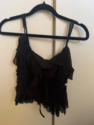Svart volang topp med tunna axelband - Snygg svart croppad topp med tunna axelband och volangdetaljer. Toppen har en luftig och lätt känsla, perfekt för att styla med höga jeans eller kjol. Materialet känns som polyester och toppen har en feminin och trendig look. Aldrig haft på mig den, prislapp är kvar.