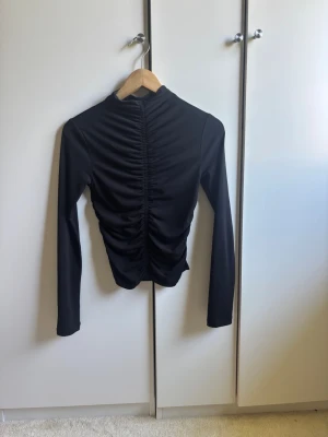 Svart långärmad topp med drapering  - Snygg svart polotopp med långa ärmar och cool rynkad draperingsdetalj fram- och baktill. Tight passform som framhäver siluetten. Jag har tyvärr vuxit ur den bara efter ett fåtal användningar. 