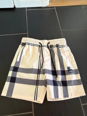 Burberry rutiga shorts - Snygga shorts från Burberry med klassiskt rutmönster i beige, svart och vitt. De har resårmidja med svart dragsko och sidofickor. Materialet är lätt och snabbtorkande, perfekt för sommaren. Burberry-logga med riddare tryckt på benet. Passar både M och L. Kom gärna med prisförslag så ska vi försöka lösa ett bra pris!