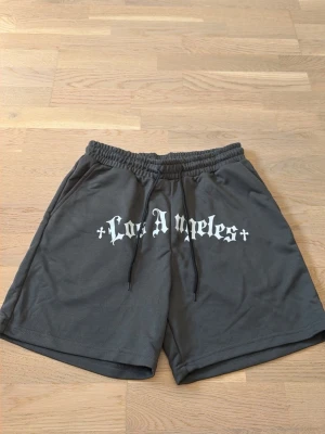 Grå shorts med Los Angeles-tryck - Mörkgrå shorts med resårmidja och snörning framtill. Stort vitt tryck med texten 'Los Angeles' i gotisk stil över framsidan. Sköna och avslappnade, perfekta för sommaren.