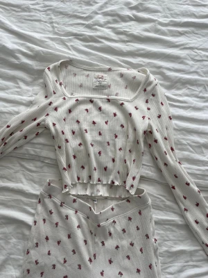 Vitt ribbat mjukisset med blommor - Säljer ett vitt ribbat set från Censored med långärmad topp och matchande underdel. Setet har små röda blommor över hela tyget, fyrkantig ringning och vågig kant nertill. Materialet är stretchigt och väldigt mjukt.