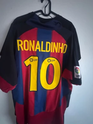 Barcelona Ronaldinho matchtröja Nike - Säljer en klassisk FC Barcelona matchtröja med Ronaldinho och nummer 10 på ryggen. Tröjan har blå och röda vertikala ränder, gul Nike-logga och LFP-märke på ärmen. Perfekt för dig som älskar fotboll och vill hylla en legend.