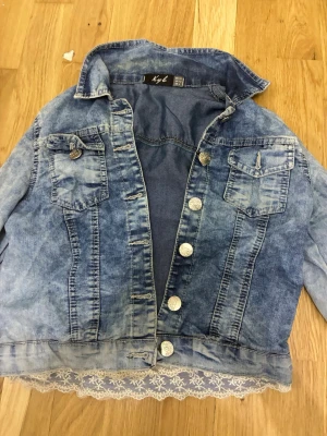 Jeansjacka med spetskant från Kyl, storlek S/XS. - Den här ljusblå jeansjackan från Kyl har en sliten look och är detaljerad med en spetskant nedtill. Sitter som storlek S/XS. 