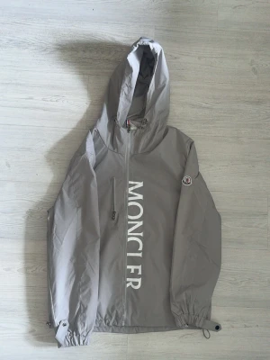 Grå vindjacka från Moncler - Snygg grå vindjacka från Moncler med stor vit logga längs dragkedjan framtill. Jackan har huva, två dragkedjeförsedda fickor och justerbara muddar. Tillverkad i lätt och tåligt material, perfekt för blåsiga dagar. Moncler-märke på ärmen ger en exklusiv touch.