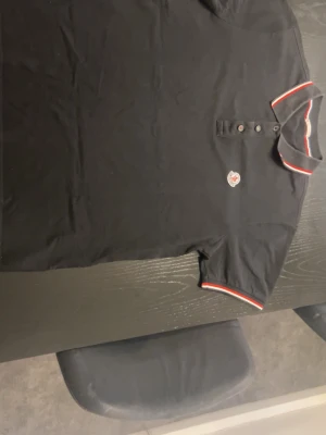 Svart pikétröja från Moncler - Svart pikétröja från Moncler med klassisk krage och knappar framtill. Tröjan har röda och vita ränder på krage och ärmslut samt Moncler-logga på bröstet. Perfekt för en stilren och sportig look.