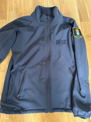Mörkblå jacka med dragkedja - Mörkblå jacka med hel dragkedja framtill och hög krage. Jackan har två sidofickor med dragkedja och ett emblem på höger ärm. Materialet känns slitstarkt och passar bra till vardags. Enkel och stilren design.
