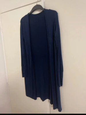 Mörkblå lång kofta utan knappar passar S-M - Lång och tunn mörkblå kofta med långa ärmar och öppen front. Koftan har minimalistisk design och små knappar vid ärmsluten. Perfekt att slänga över en t-shirt eller topp för en avslappnad look.