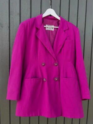 Vintage Fuchsia kavaj från Flor da Moda - Stilren fuchsiafärgad kavaj från Flor da Moda med dubbelknäppning och dekorativa knappar. Klassisk krage, två framfickor och en bröstficka. Tillverkad i ett slätt tyg som ger en snygg siluett. Perfekt för dig som vill sticka ut med färg.