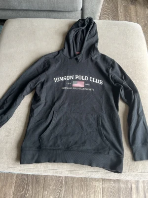 Svart hoodie från Vinson Polo Club - Cool svart hoodie från Vinson Polo Club med tryck på bröstet och amerikansk flagga. Klassisk huva och stor magficka framtill. Materialet är mjukt och bekvämt, perfekt för att chilla eller hänga med kompisar. Snygga detaljer med röd söm på insidan av kragen.