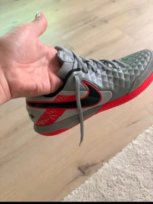 Nike grå/röda fotbollsskor - Säljer ett par grå Nike fotbollsskor med röd sula och svarta detaljer. Skorna har ett mönstrat material på ovandelen för bättre bollkontroll och klassisk snörning. Perfekta för dig som vill ha stilrena och funktionella skor på planen.
