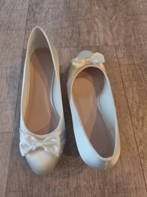 Satinskor med kilklack och rosett st 39 - Snygga  ballerinaskor med liten kilklack och dekorativ rosett framtill. Färg Ivory  Skorna har rundad tå och är tillverkade i lyxigt satin som ger en elegant look. Perfekta för dig som vill ha något stilrent och klassiskt.
