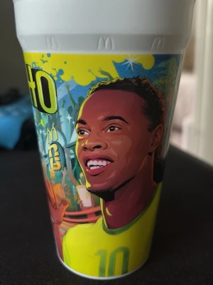 McDonald's FIFA plastmugg - Cool plastmugg från McDonald's med färgglad FIFA-design. Muggen har illustrationer av fotbollsspelare, nummer och detaljer i gult, blått, grönt och rött. Perfekt för samlare eller dig som gillar unika dryckesbehållare med sporttema.