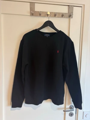 Svart sweatshirt från Polo Ralph Lauren - Klassisk svart sweatshirt från Polo Ralph Lauren med rund halsringning och den ikoniska röda logotypen broderad på bröstet. Tröjan har långa ärmar och ribbade muddar vid ärmslut och nederkant. Står XL som storlek men det är Medium size.