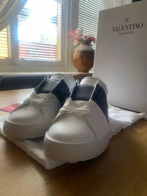 Valentino Garavani vita sneakers - Stilrena vita sneakers från Valentino Garavani med bred svart läderdetalj över snörningen. Klassisk låg modell med rund tå och vita skosnören. Tillverkade i slätt läder för en lyxig känsla. Perfekta för dig som gillar exklusiv och modern design.