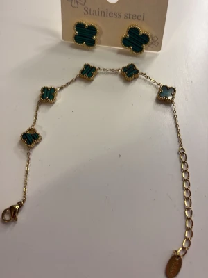 Guldfärgat armband med gröna klöver - Snyggt armband i guldfärgad stainless steel med fem fyrklöverformade detaljer i grön sten och guldkant. Justerbar längd med karbinlås och liten bricka vid låset. Perfekt accessoar för att lyfta din stil. Saknar en del av på en av blommorna men syns inte från långt håll.