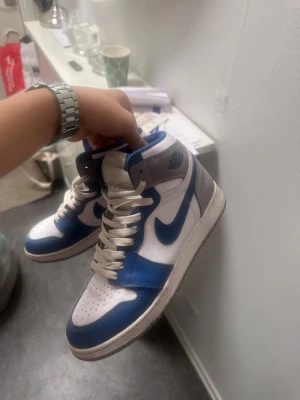 Nike Air Jordan 1 High blå/vit - Säljer ett par Nike Air Jordan 1 High sneakers i blått, vitt och grått.dem är 7 månader gamla men har in nye används vid vintern alls pågrund av mockan på skon. Denna typen av skor är Perfekt för dig som gillar streetwear. Finns med skolåda.det finns smuts på skon fast inget man inte kan göra rent.