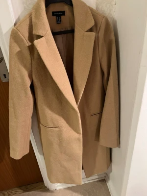 Beige kappa  - Stilren beige kappa  med klassisk krage och raka linjer. Kappan har två fickor framtill och är helfodrad med ett mjukt innerfoder. Perfekt för dig som gillar en enkel och tidlös look.