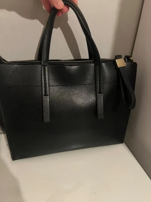 Svart stilren handväska i skinnimitation - Snygg svart handväska i skinnimitation med raka linjer och två handtag. Väskan har guldfärgade metalldetaljer och dragkedja upptill. Perfekt för dig som gillar minimalistisk och modern design.