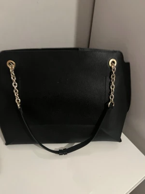 Svart handväska med kedjedetaljer - Stilren svart handväska i konstläder med guldfärgade kedjedetaljer på axelremmarna. Väskan har en enkel, rektangulär form och passar perfekt för dig som gillar en klassisk look med en twist. Rymmer allt du behöver till vardagen och har snygga metallringar som detaljer.