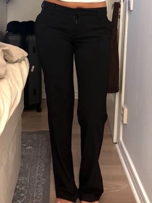 Svarta kostymbyxor med raka ben - Snygga svarta kostymbyxor med raka ben och låg midja. Byxorna har bälteshällor, dragkedja och knapp framtill. Perfekta för en clean och stilren look. Materialet ser ut att vara en mjuk och stretchig blandning som sitter bekvämt.