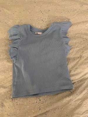 Blå ribbad topp med volangärmar Zara - Supersöt ljusblå ribbad topp från Zara med kort, croppad passform och volangdetaljer på axlarna. Rund halsringning och stretchigt material som sitter snyggt. Perfekt för dig som vill ha en trendig och bekväm topp till vardagslooken.