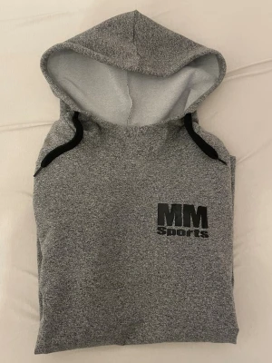 Grå hoodie från MM Sports - Säljer en grå hoodie från MM Sports med svart tryck både fram och bak. Hoodien har huva med svarta snören och är tillverkad i mjukt sweatshirtmaterial. Perfekt för gymmet eller chill dagar. Klassisk passform och lång ärm.