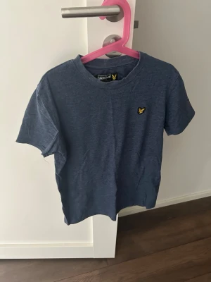 Blå t-shirt från Lyle & Scott - Snygg blå t-shirt från Lyle & Scott med klassisk rund hals och korta ärmar. T-shirten har en gul broderad logga på bröstet och är tillverkad i mjuk bomull. Perfekt för en avslappnad och stilren look.