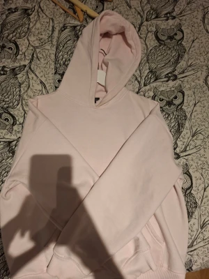 Ljusrosa hoodie med huva - Säljer en stilren ljusrosa hoodie med klassisk huva och känguruficka framtill. Perfekt för dig som gillar en enkel och clean look. Mjuk bomullskänsla och ribbade muddar vid ärmslut och nederkant.