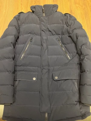 Svart Jerem pufferjacka med dragkedjor - Snygg svart pufferjacka med hög krage och två snedställda bröstfickor med dragkedja samt två stora fickor med lock och tryckknappar. Jackan har quiltad design och dragkedja framtill. Perfekt för kalla dagar och ger en stilren look. 