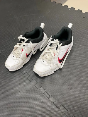 Nike vita sneakers med röd swoosh - Fräscha vita sneakers från Nike med klassisk röd swoosh på sidorna och svarta detaljer. Skorna har lågt skaft, svart innersula och vit ovandel i läder med meshpartier. Perfekta för en sportig och clean look.
