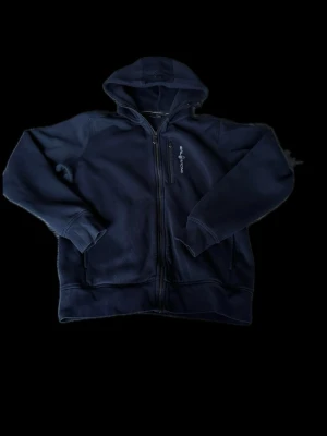 Mörkblå zip-hoodie från Sail racing - Mörkblå hoodie med dragkedja från Sail racing. Klassisk modell med huva, två fickor fram och diskret logga på bröstet. Tillverkad i mjukt material som passar perfekt för en avslappnad stil. Snygg och enkel att matcha med det mesta. Strl 164/170