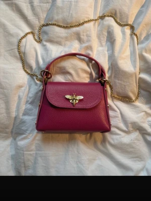 Mörkrosa handväska med guldkedja - Snygg mörkrosa handväska i läder med guldfärgad kedja och handtag. Väskan har ett dekorativt guldfärgat bi-spänne på framsidan och en stilren, boxig form. Perfekt accessoar för att lyfta din outfit. Väskan är tillverkad och såld i frankrike och är i äkta läder 