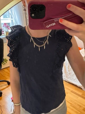 Mörkblå blus med volangärmar - Supersöt mörkblå blus med broderade volangärmar. Toppen har rund halsringning och är ärmlös med breda, volangiga axelpartier som ger en romantisk vibe. Perfekt att styla med smycken för en trendig look.