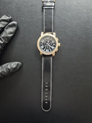 Guldaktig Klocka - Stilren klocka från Deni & Gabi med svart läderarmband och vita sömmar. Urtavlan är svart med stora vita siffror och tre dekorativa små urtavlor. Runt boetten sitter glittrande stenar och boetten är i guldton. Har man fler frågor så är det bara att fråga.