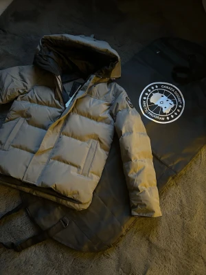 Canada gose macmillan  - Snygg grå pufferjacka från Canada Goose med svart innerfoder och huva. Jackan har klassisk patch-logga på ärmen och raka, quiltade paneler. Perfekt för kalla dagar och riktigt varm. Dragkedja framtill och två sidofickor.