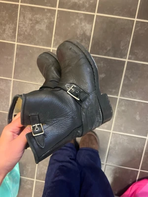 Svarta läderstövletter med spännen - Svarta boots i äkta läder med två silverfärgade spännen på sidan. Klassisk rund tå och låg klack. Snyggt robusta och passar perfekt till jeans eller kjol. Tidlös design med kort skaft och dragkedja på insidan.