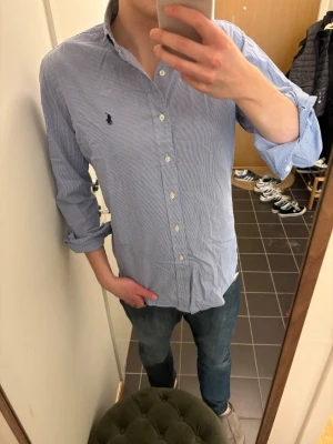 Blårutig skjorta från Ralph Lauren - Klassisk blårutig skjorta från Ralph Lauren med button-down krage och lång ärm. Skjortan har broderad logga på bröstet och vita knappar framtill. Tillverkad i mjuk bomull och har en avslappnad passform.